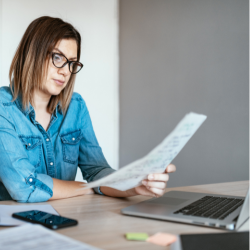Serious young woman in glasses and a denim shirt sitting at a laptop looking at a piece of paper